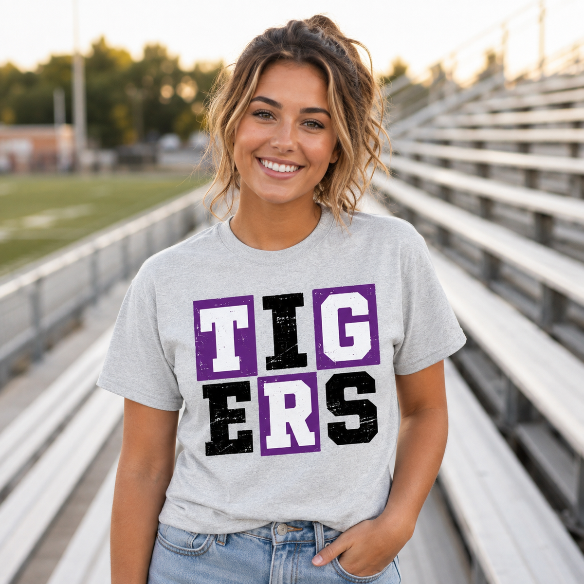 Model wearing ash gray Tigers purple and black grid graphic tee at football bleachers